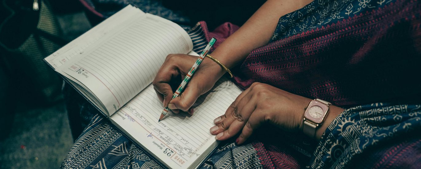 Person writing in a notebook with a patterned pen, wearing a colorful patterned outfit.